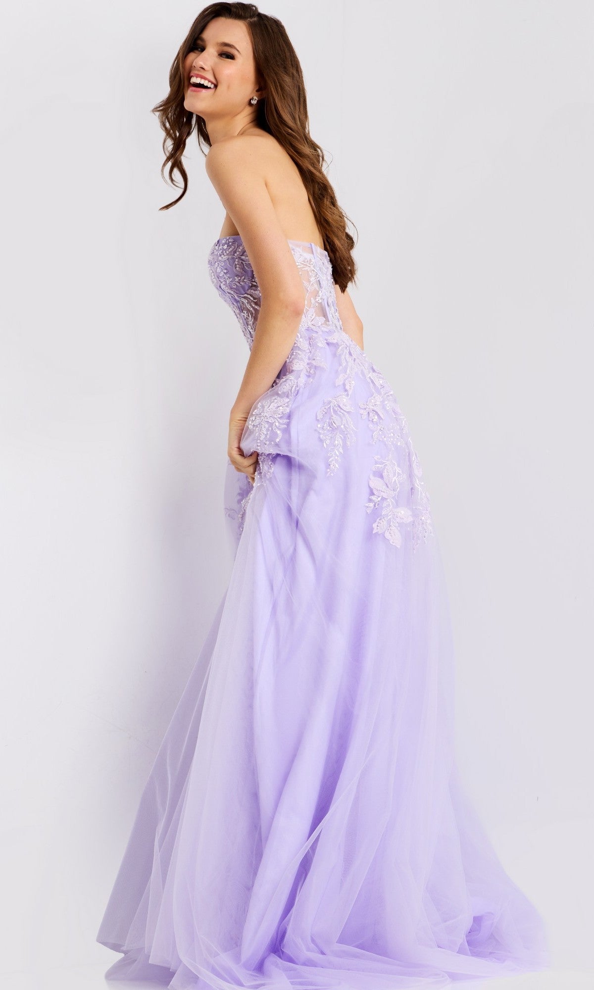 A woman models the Jovani 49656, a strapless lavender lace long prom dress with a sweetheart neckline, smiling as she stands sideways against a plain white background.